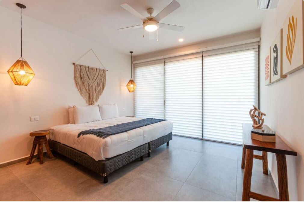 Ein Schlafzimmer mit Bett, Deckenventilator, Wandkunst aus Makramee und einem Fenster mit Jalousien.