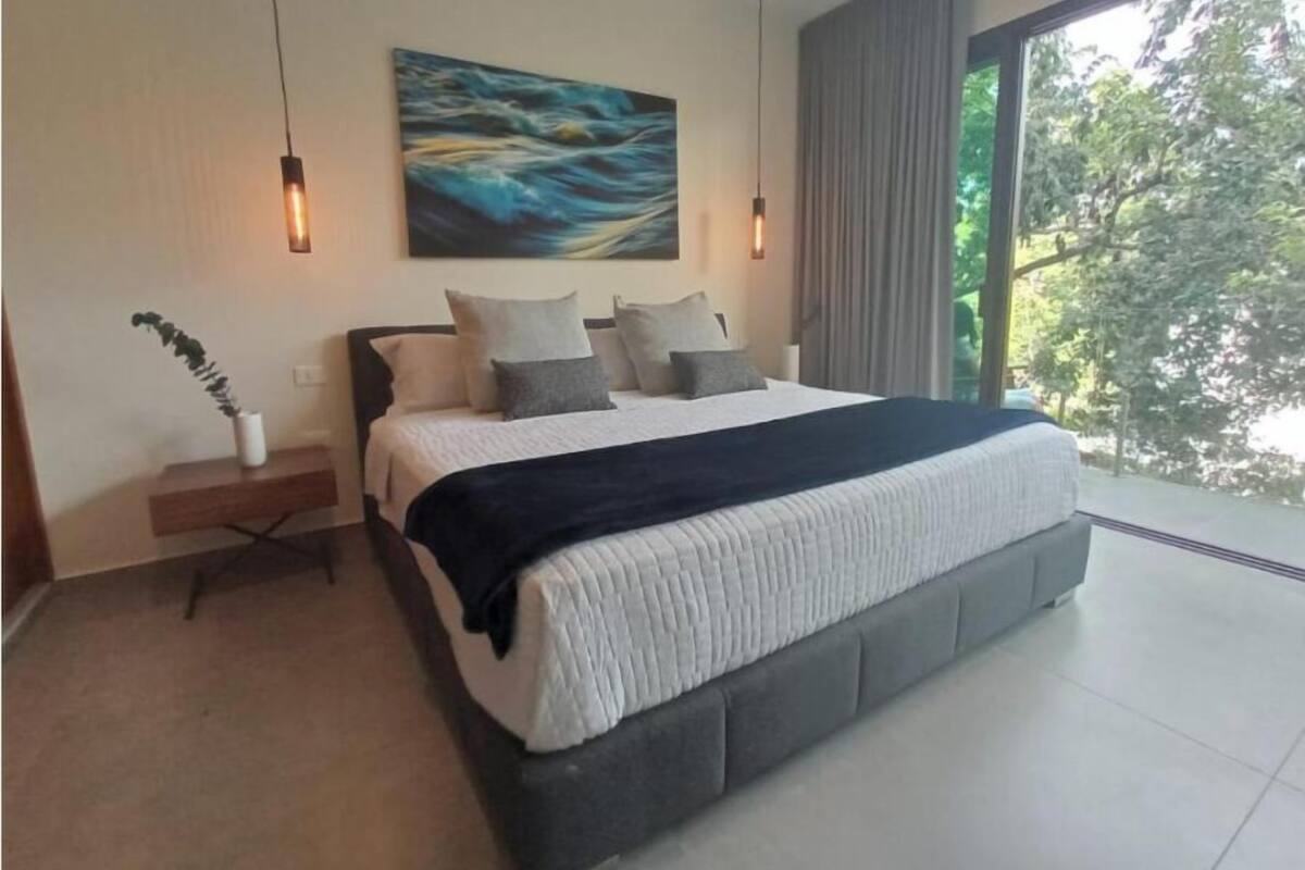 Un dormitorio moderno con una cama grande, un cuadro en la pared y vistas a árboles a través de una ventana.