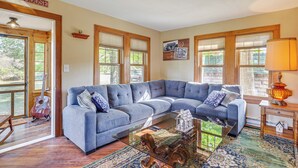 A spacious living room with a large sectional sofa, a glass coffee table, and a wooden floor. There are large windows with blinds, a guitar leaning against the wall, and a sign that reads "FARMHOUSE". - Pellston Farmhouse w/ Snowmobile Trail Access! (Pellston)