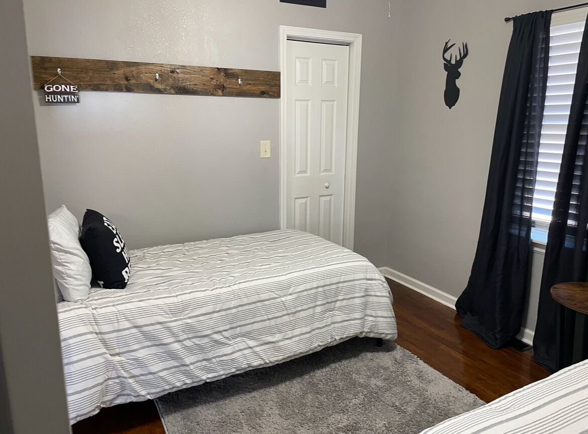 Une chambre à coucher avec un lit, une porte, une tête de cerf accrochée au mur, un porte-manteau et une fenêtre avec des rideaux.