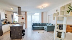 Eine moderne Küche mit Essbereich, ein Wohnzimmer mit Ecksofa und ein weißes Regal mit Pflanzen.