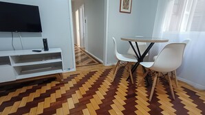 A room with a flat-screen TV on a white stand, a small table with two white chairs, and a patterned wooden floor. - Cozy apartment, great location with garage (Centro Histórico Porto Alegre)