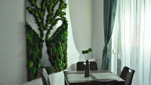 A dining area with a table and chairs, a wall-mounted moss art piece, and a window with curtains. - Mirvana Apartments Metheor Executive Suite (Timișoara)