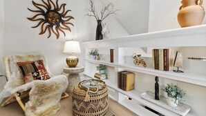 A cozy living room with a white armchair, a round woven ottoman, a sun-shaped wall decoration, and a bookshelf with books and decorative items. - Lee Martinez Loft Near Poudre Trail and Old Town (Fort Collins)