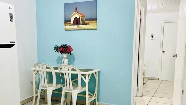 Un pequeño comedor con una mesa y sillas blancas, un jarrón con flores y un cuadro en la pared.