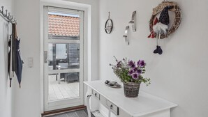 A clean, modern interior with a white wall, a door leading to a balcony, a small white table with a potted plant, and a welcome wreath. - Awesome home in Hirtshals with sauna (Hirtshals)