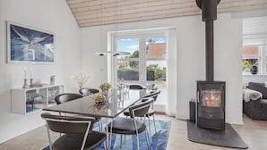 A modern living room with a glass dining table, black chairs, a black wood-burning stove, and a large window with a view of the outdoors. - Awesome home in Hirtshals with sauna (Hirtshals)