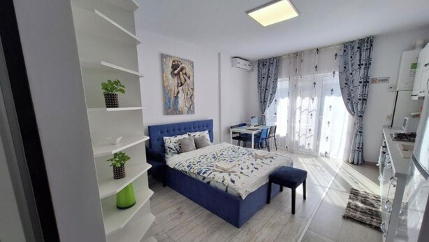 A modern bedroom with a large bed, a desk, and a chair. There are shelves with plants and a painting on the wall. - Mirvana Apartments Allure Studio (Timisoara)