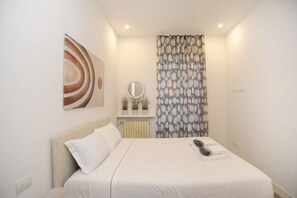 Un dormitorio con cama, mesita de noche con espejo, radiador y cortina estampada con hojas.