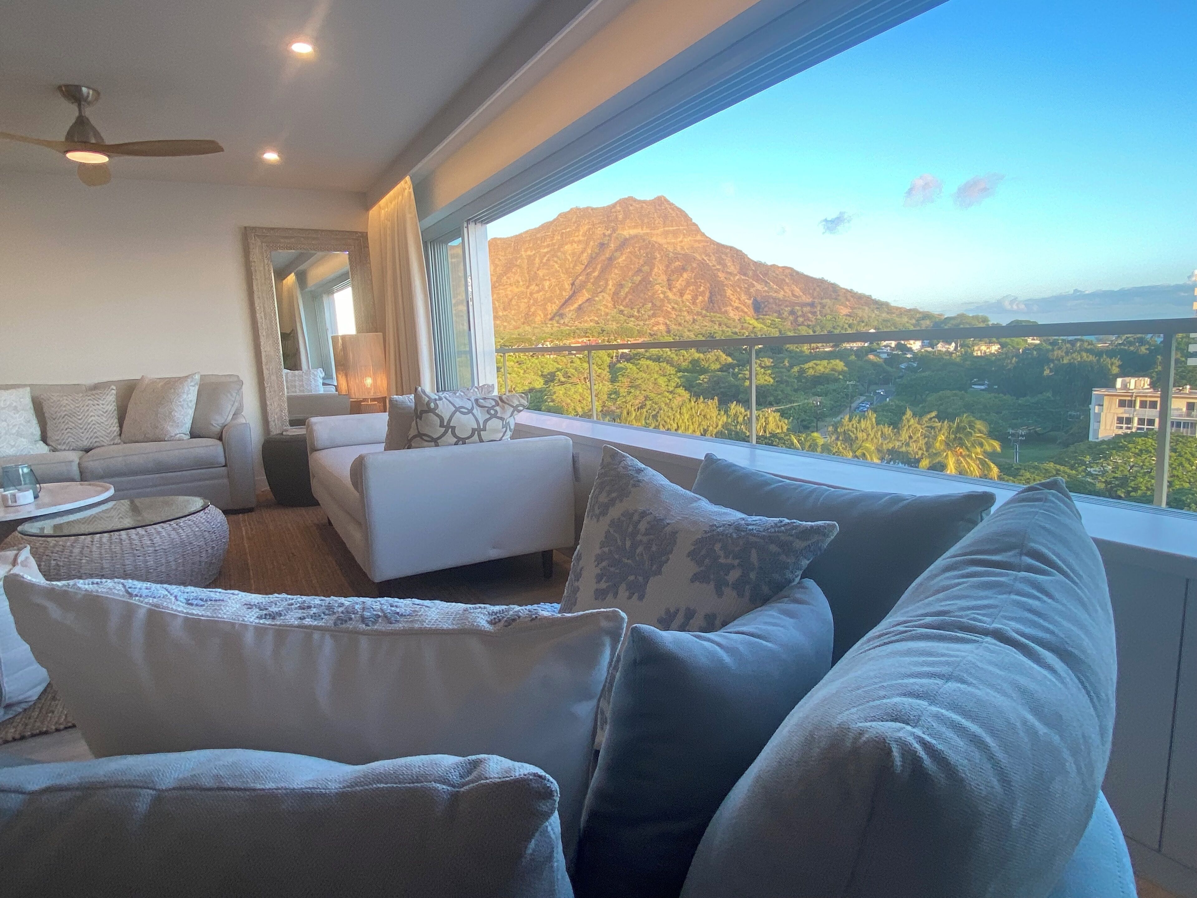 Ein modernes Wohnzimmer mit einer Couch, Sesseln und einem Glastisch. Ein großes Fenster bietet Blick auf Berge und Grünflächen.