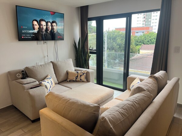 A living room with a large flat-screen TV mounted on the wall, a beige sofa, and a window with a view of buildings and trees. - Entire apartment in zone 10! (Ciudad de Guatemala)