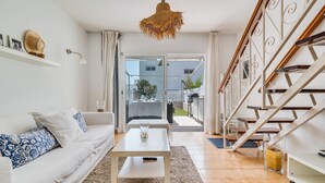Ein modernes Wohnzimmer mit einem weißen Sofa, einem Couchtisch und einer Pendelleuchte. Es gibt eine Treppe mit Geländer und einen Blick nach draußen.