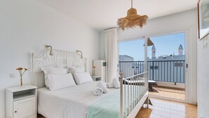 Ein Schlafzimmer mit einem Bett, Nachttischen, einer Hängeleuchte und einem Balkon mit Blick auf Gebäude.