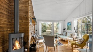 Um quarto aconchegante com lareira a lenha, um sofá, cadeiras e janelas amplas com vista para o exterior.