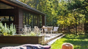 Uma cabana de madeira com área de varanda, com cadeiras e mesa brancas, rodeada por um jardim com diversas plantas e uma cerca de madeira.