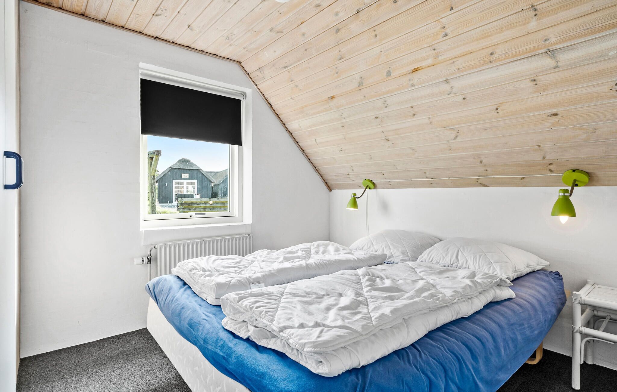 Un dormitorio con techo inclinado, una ventana con vista a un edificio, una cama con ropa de cama blanca y azul, y dos lámparas de pared verdes.