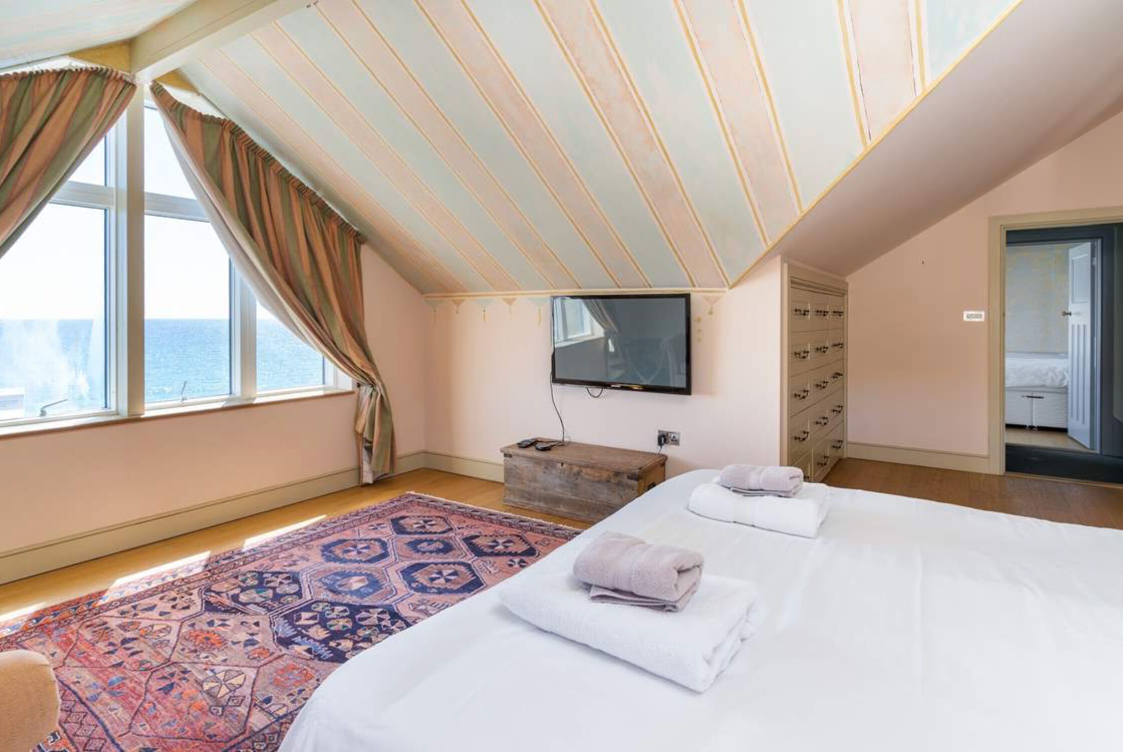 A bedroom with a large bed, a TV mounted on the wall, a wooden chest of drawers, and a view of the sea through a window.