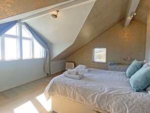 A bedroom with a sloped ceiling, a bed with white linens, a radiator, a window with curtains, and a small bedside table with a vase of flowers. - Private Cliff top Retreat by Perranuthnoe Beach (Penzance)