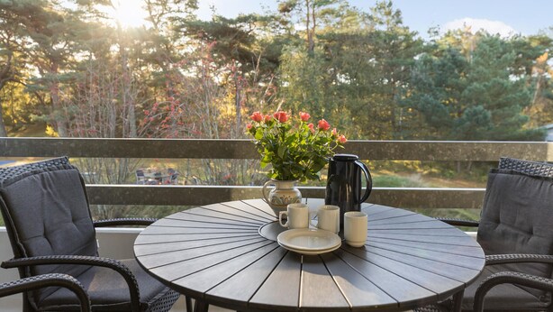 A round wooden table with a vase of pink flowers, a coffee pot, and white mugs on a balcony with wicker chairs. - Nice apartment in Nexø with WiFi (Nexø)