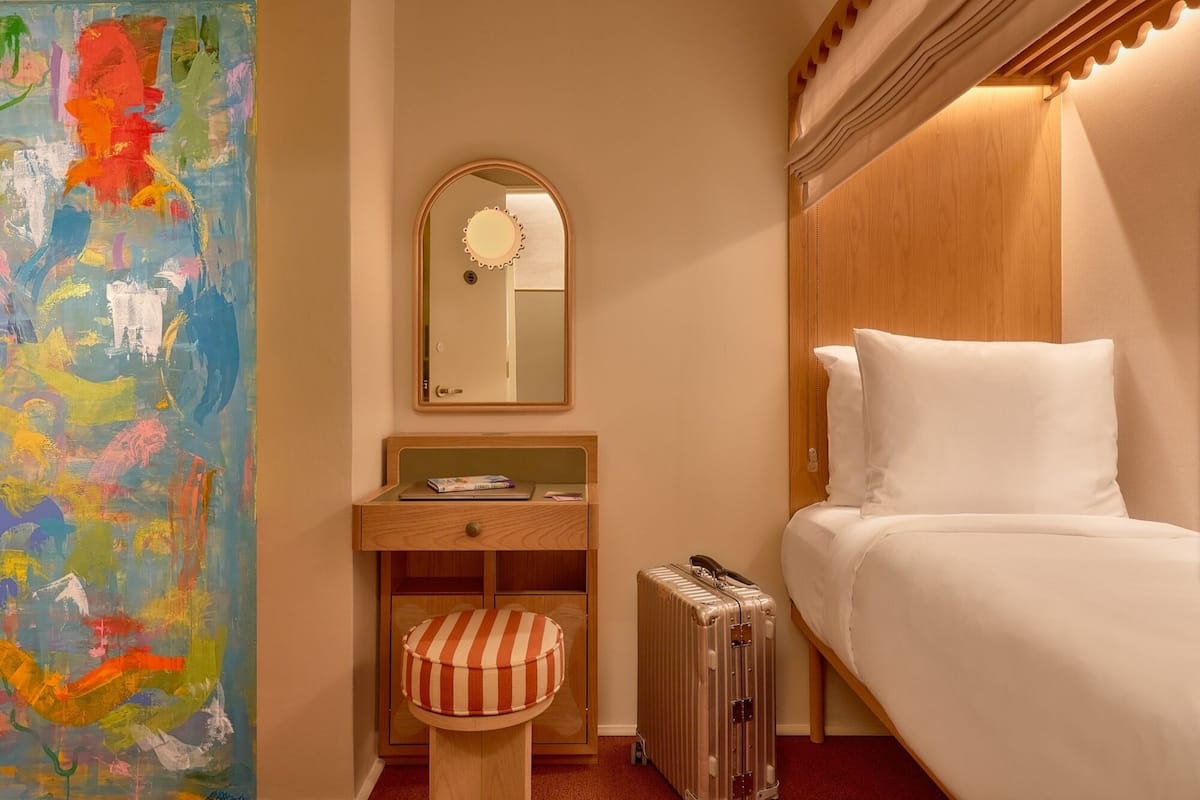 A hotel room with a bed, a wooden nightstand with a lamp, a striped ottoman, a suitcase, and a colorful abstract painting on the wall.
