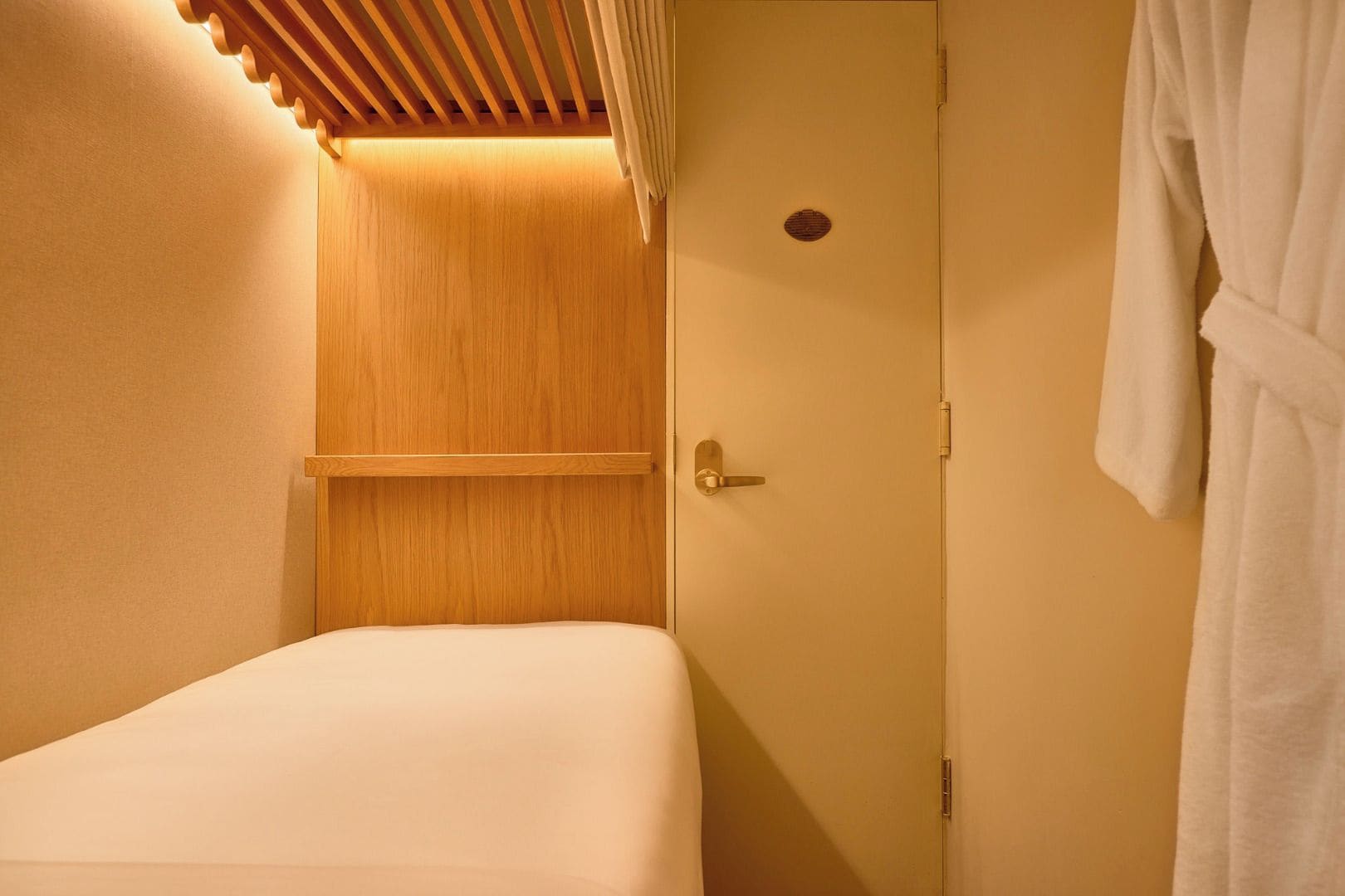 A small, single-bed room with a wooden panel wall, a door with a round window, and a white bathrobe hanging on the right.