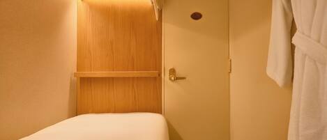 A small, single-bed room with a wooden panel wall, a door with a round window, and a white bathrobe hanging on the right.