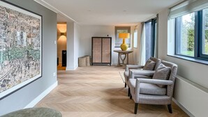 Una habitación amplia con piso de madera en patrón de espiga, un sillón tapizado gris, una mesita de madera redonda con una lámpara amarilla y una obra de arte grande enmarcada en la pared.