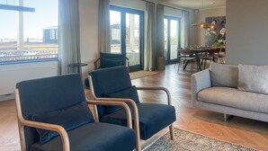 Amplia sala de estar con sofá, dos sillones, una mesa de comedor con sillas y ventanas grandes con cortinas.