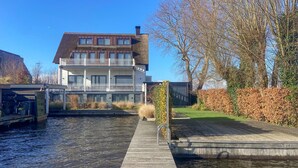 Un edificio moderno de dos pisos con un balcón, un muelle de madera que se adentra en un canal, y árboles con hojas al fondo.