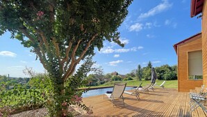 Terraza de madera con sillones de descanso con vista a una piscina y un paisaje pintoresco de un lago y colinas.