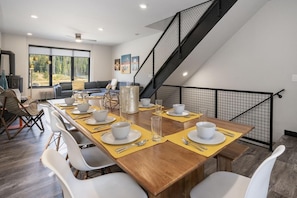 Comedor con una larga mesa de madera dispuesta para cuatro personas, sillas blancas y una escalera de metal negro.