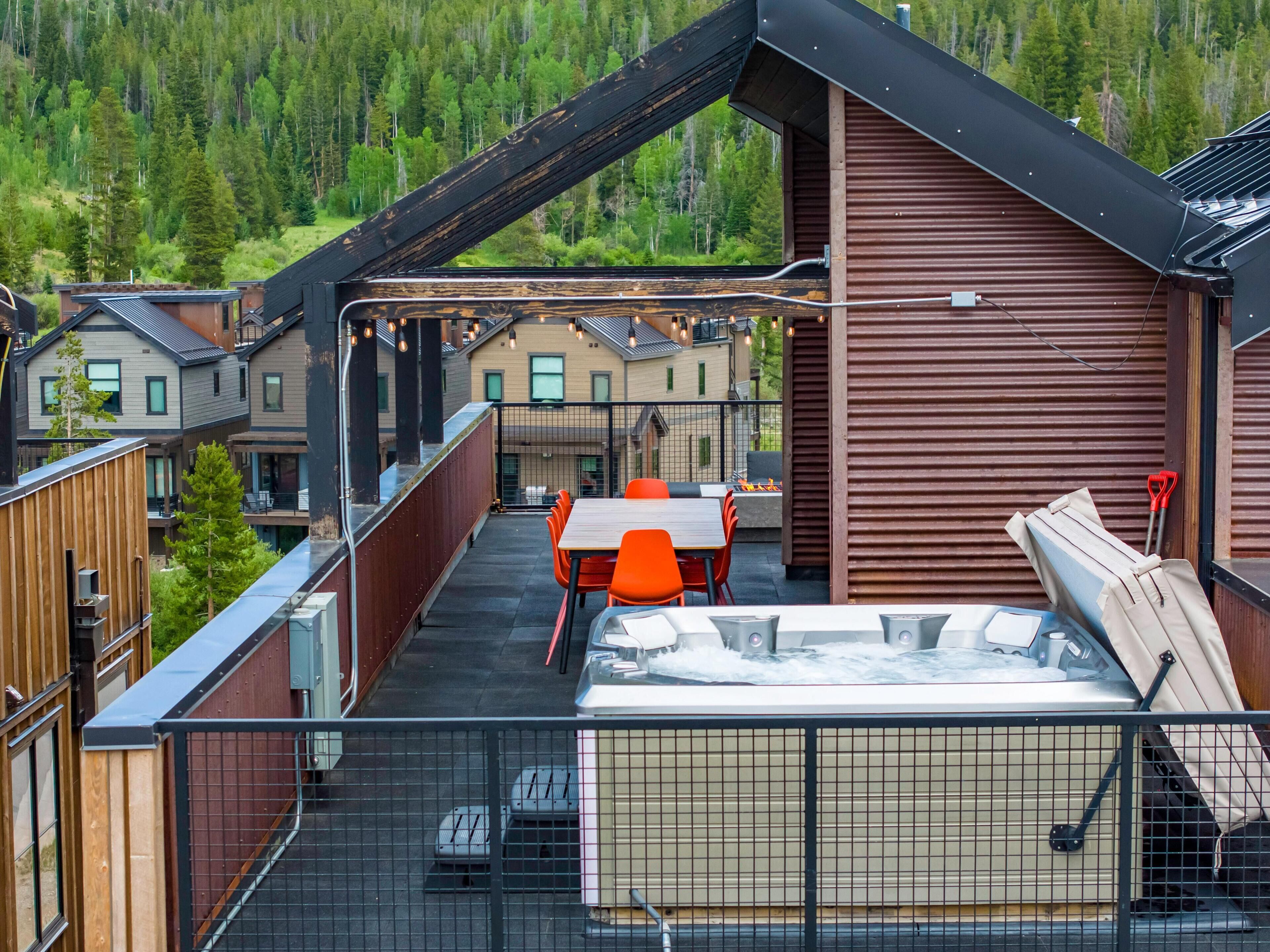 A rooftop terrace with a hot tub, outdoor seating, and a view of a forested area.