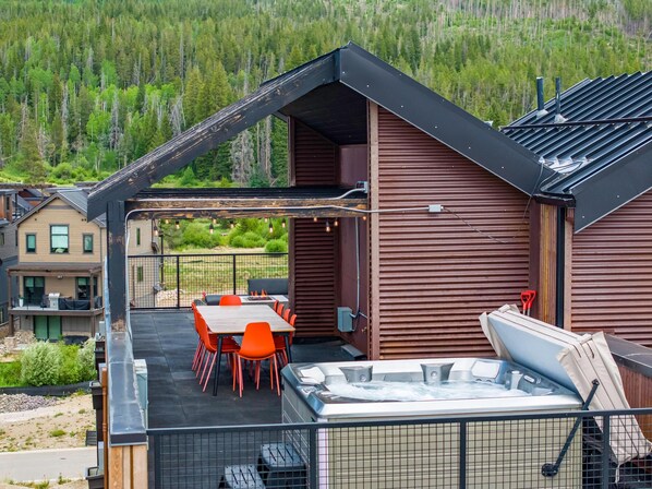 A modern outdoor patio with a hot tub, a dining table with chairs, and a view of a forested area. - Row 41 | 4BR, Rooftop Hot Tub, Walk to Town (Winter Park)