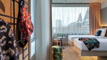 Ein Hotelzimmer mit Bett, einem kleinen Tisch, einem Koffer und Blick auf Gebäude durch durchsichtige Vorhänge.