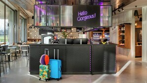 A modern bar area with a neon sign reading "Hello Gorgeous!" and a bar counter with various bottles and a plant. There are chairs and tables in the foreground, and a suitcase is visible on the floor. - Moxy Antwerp (Antwerp)