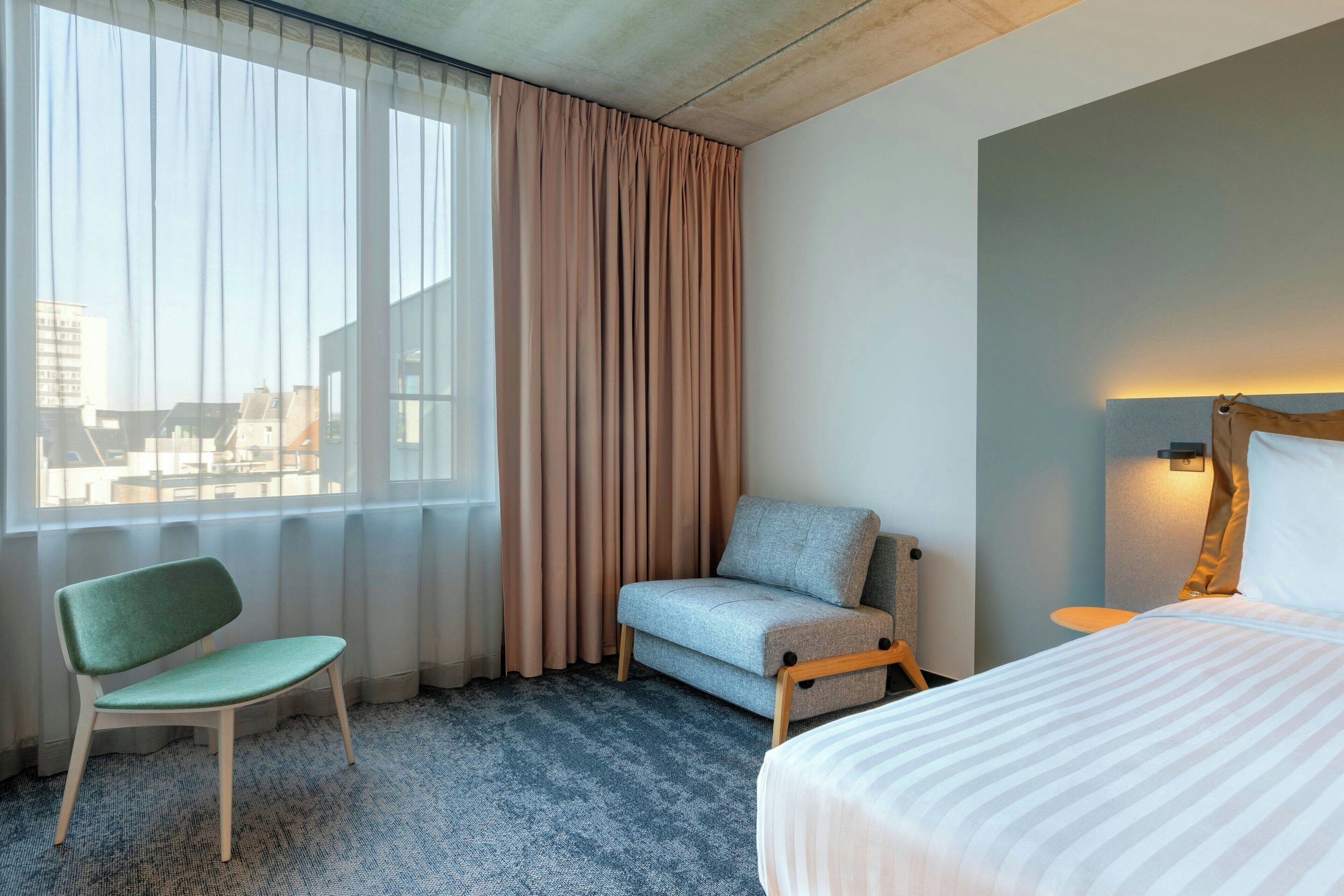 Habitación de hotel con cama, dos sillas, ventana con cortinas y vista a la ciudad.
