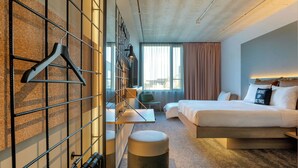 A hotel room with a bed, a desk, a chair, a hanging coat rack, and a round ottoman. - Moxy Antwerp (Antwerp)