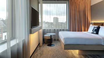 Habitación de hotel con una cama, televisión montada en la pared, una mesita redonda y una ventana con cortinas.