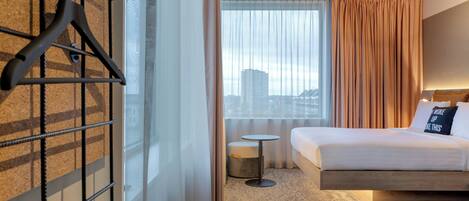 A hotel room with a bed, a small round table, a chair, a hanging coat rack, and a window with curtains.