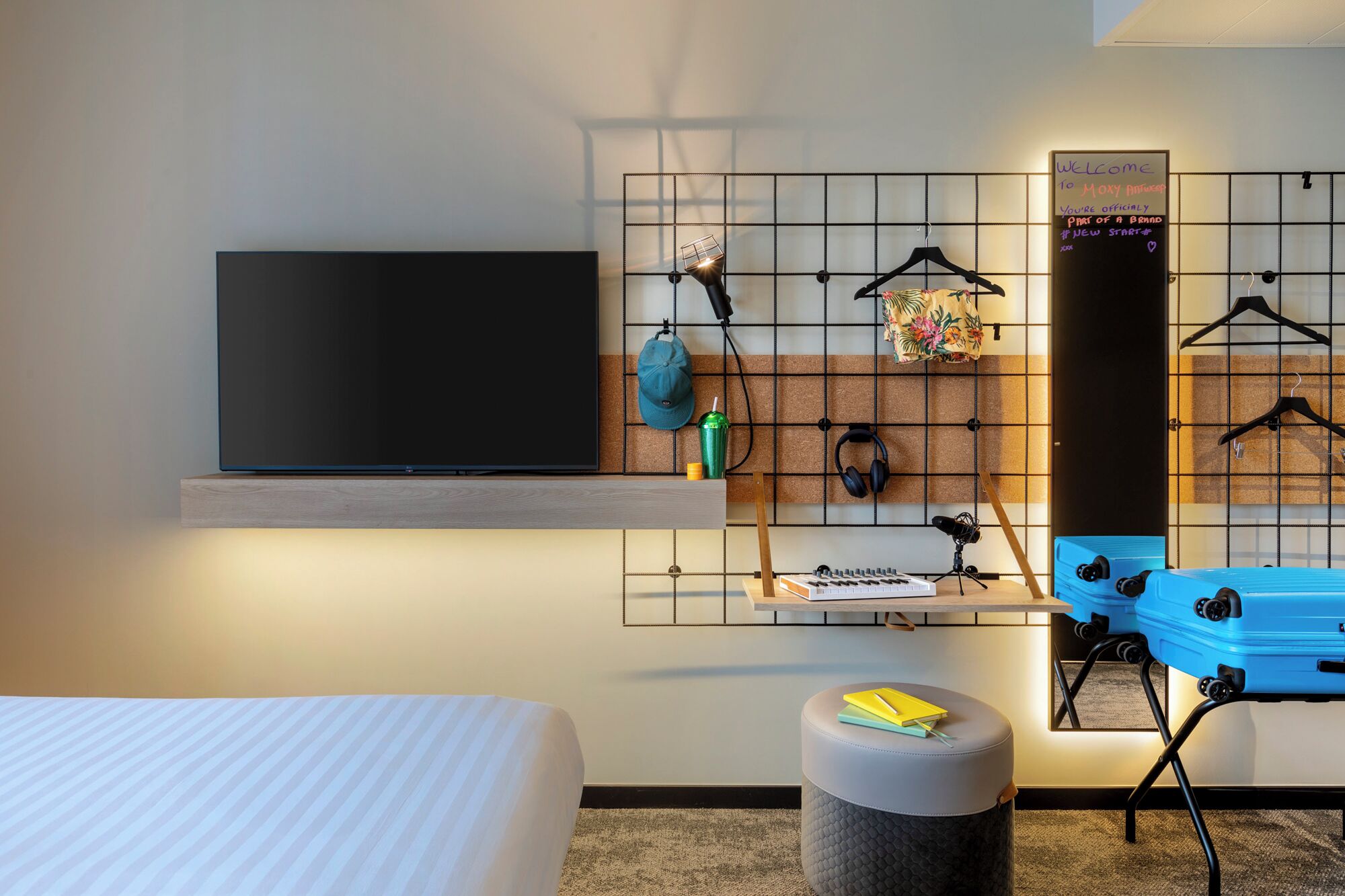 Une chambre d’hôtel avec un lit, un téléviseur fixé au mur, un bureau avec une chaise, un petit tabouret, un porte-manteau mural, une valise et un tableau noir avec un message de bienvenue.