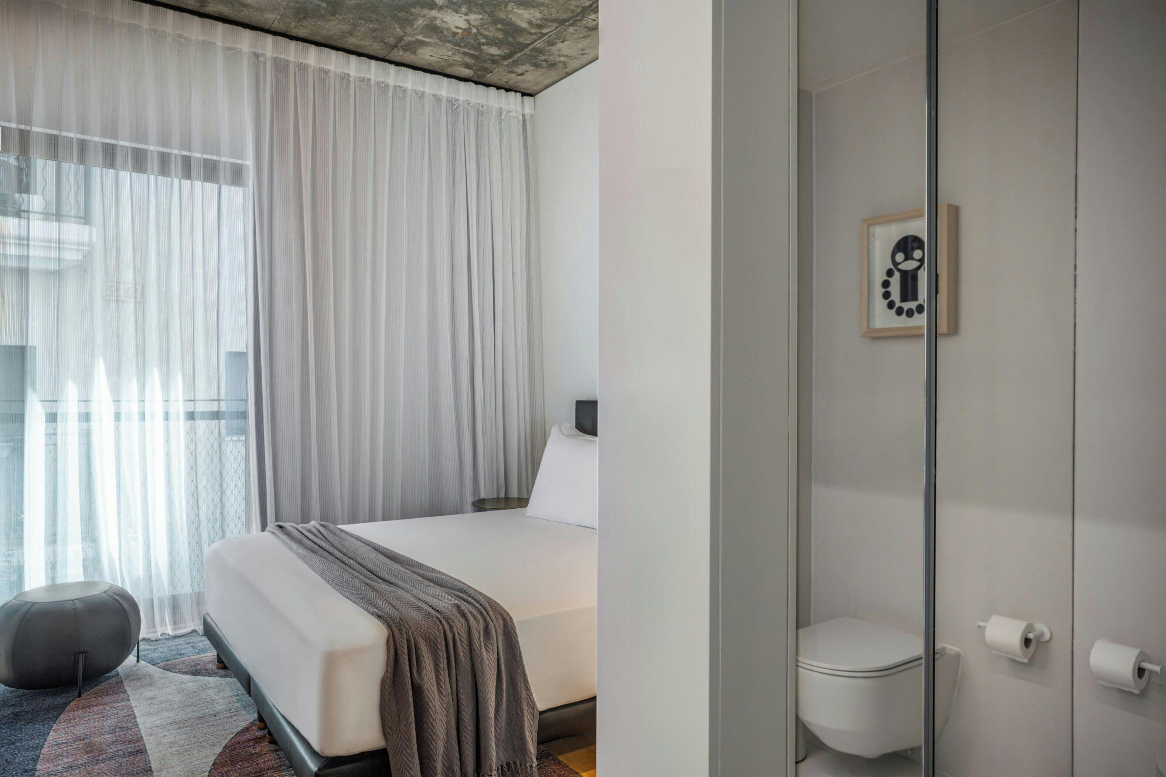 Ett modernt hotellrum med en stor säng, en duschkabin med glasväggar och utsikt över staden genom tunna, genomskinliga gardiner.