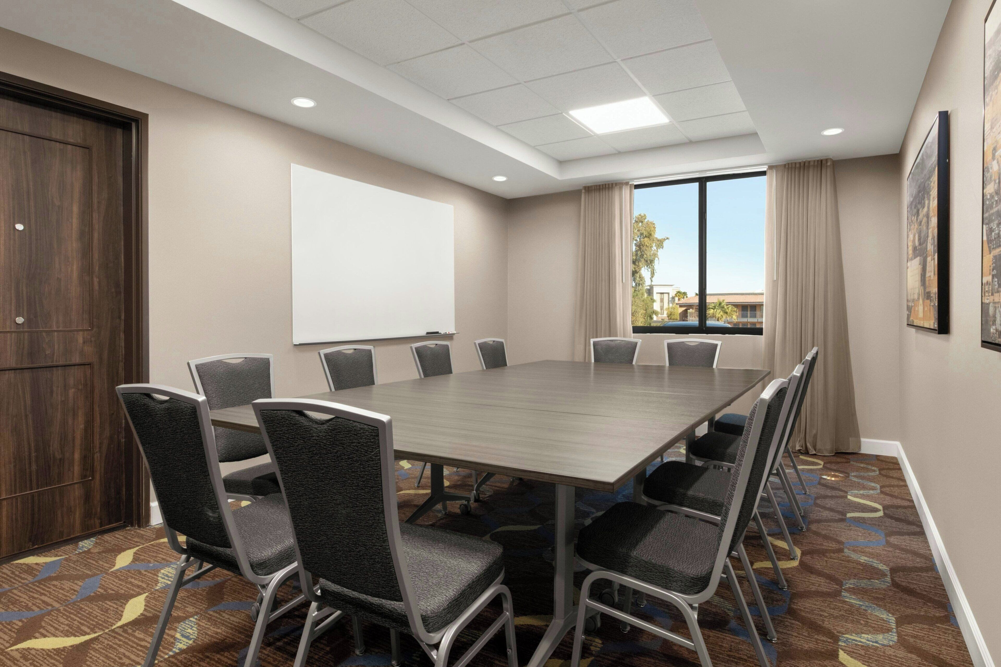 A conference room with a large table, chairs, a projector screen, and a window with curtains.