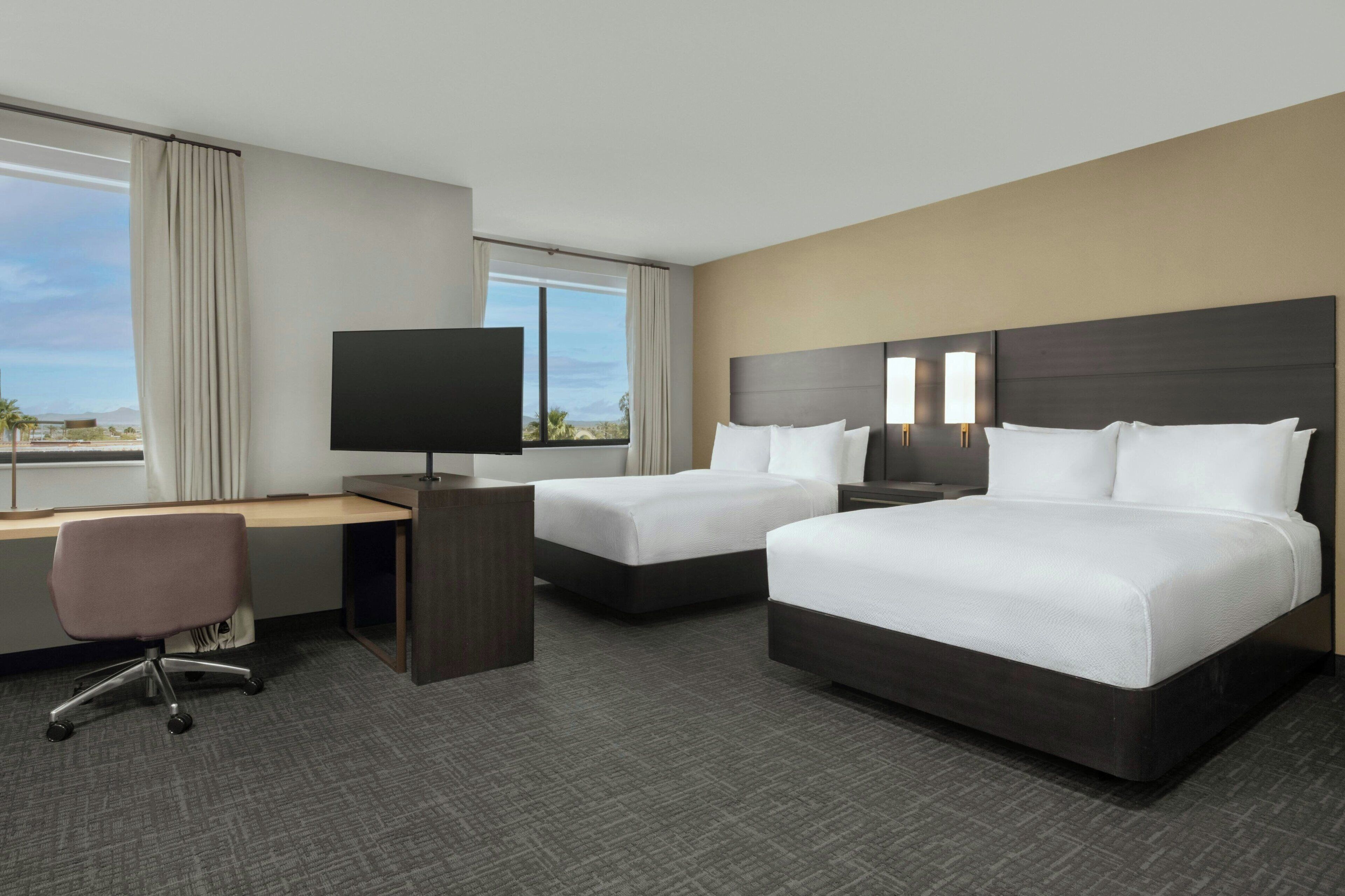 A hotel room with two beds, a desk, a chair, a TV, and a view of the outdoors.
