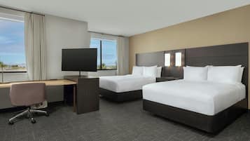 A hotel room with two beds, a desk, a chair, a TV, and a view of the outdoors.