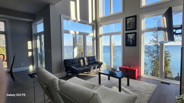 Ein modernes Wohnzimmer mit einer Couch, einem Couchtisch und einem roten Hocker. Große Fenster bieten einen Ausblick ins Freie.