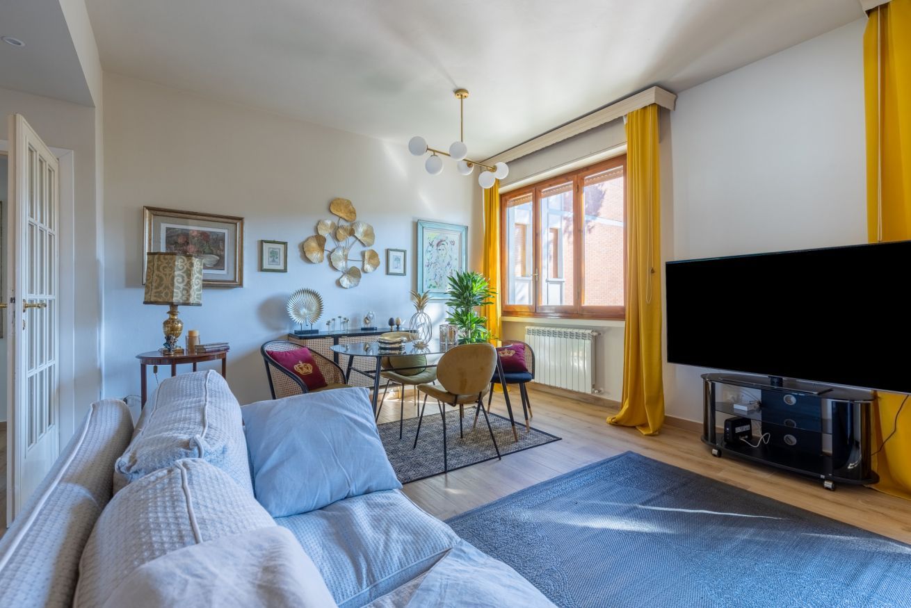 Una sala de estar con sofá, una mesa de comedor con sillas, televisor y ventana con cortinas.