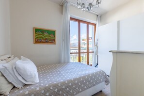 A bedroom with a bed, pillows, a window with curtains, a painting on the wall, and a balcony with a railing.