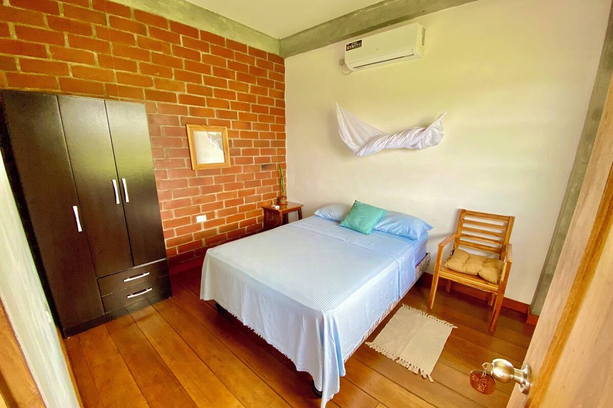 A bedroom with a brick wall, a bed with a blue bedspread, a wooden chair, a wooden wardrobe, and a white air conditioning unit.