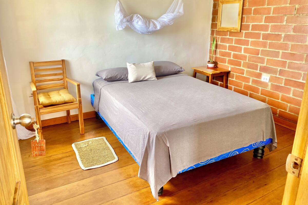 A bedroom with a bed, a wooden chair, a small table, a hanging light, and a brick wall.