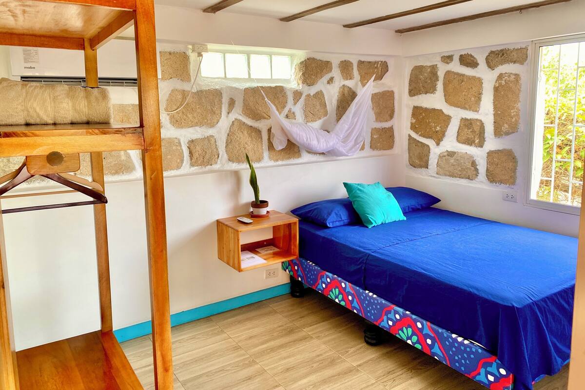 A room with a bunk bed, a small bedside table with a plant, a window with a view of greenery, and a stone wall.
